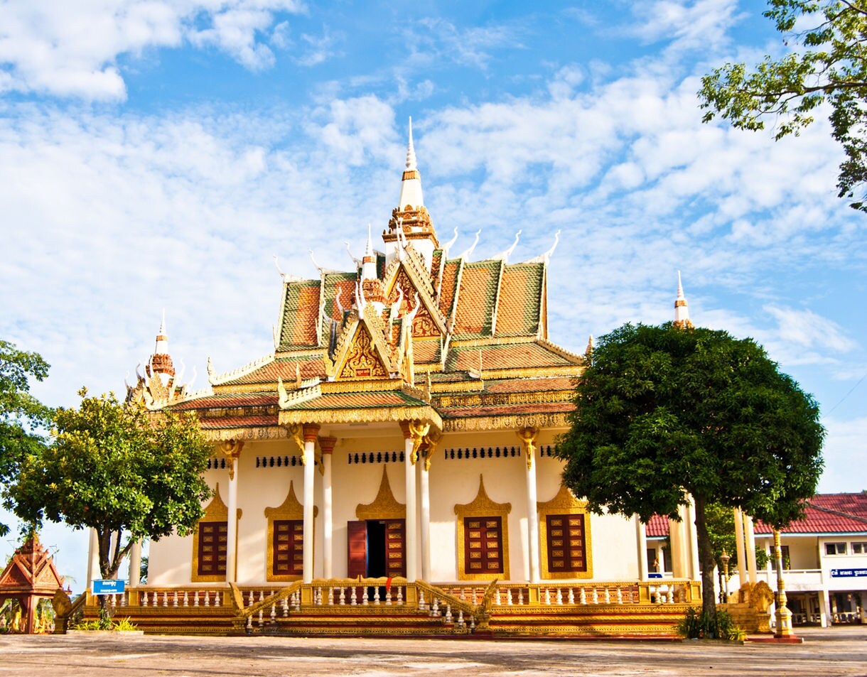 Ornate Cambodian temple with golden trim, tiered roofs and decorative spires surrounded by trees under a partly cloudy sky.
