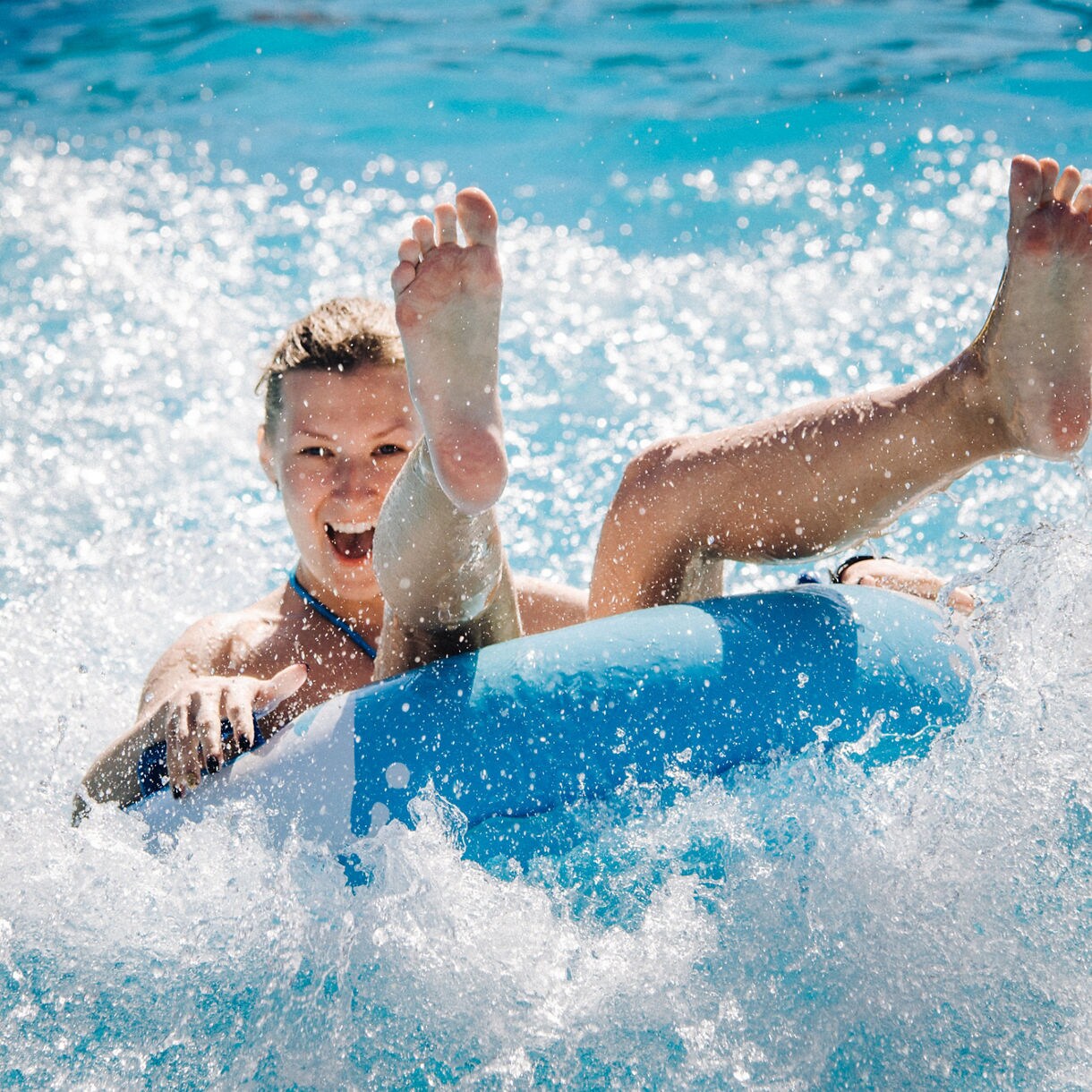 Person laughing while riding a blue inflatable tube through splashing water at a waterpark.