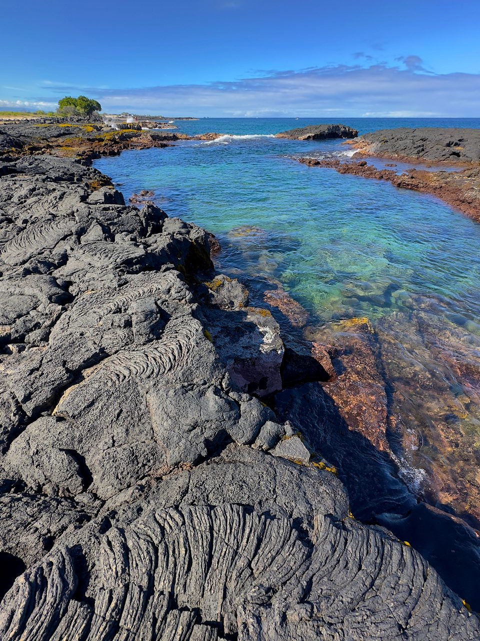 Clear blue tide pool surrounded by rugged black lava rock formations at Wawaloli Beach Park under a bright sky.
