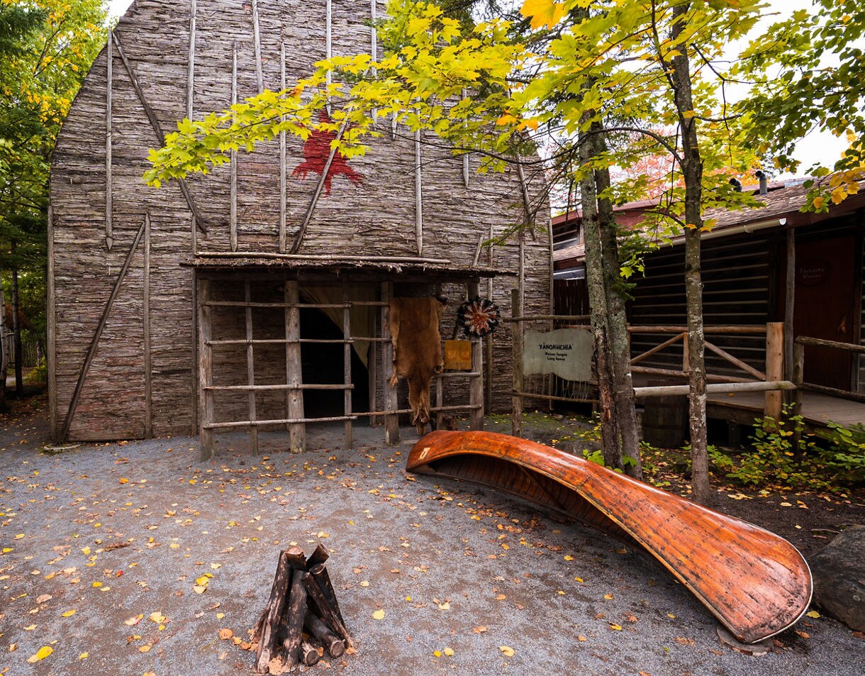 A reconstructed Huron-Wendat longhouse in Wendake, surrounded by autumn trees, with a wooden canoe, fire pit and traditional tools displayed outside.