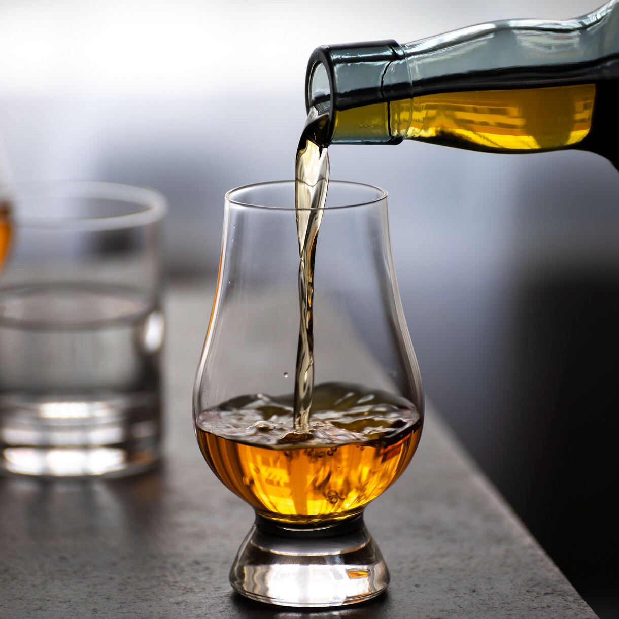 Close-up of whisky being poured from a bottle into a tulip-shaped glass, with two other glasses—one of whisky and one of water—blurred in the background.