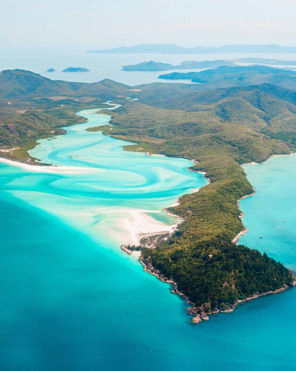 Aerial view of Whitehaven Beach showing bright turquoise water curving around forested islands in the Whitsundays.