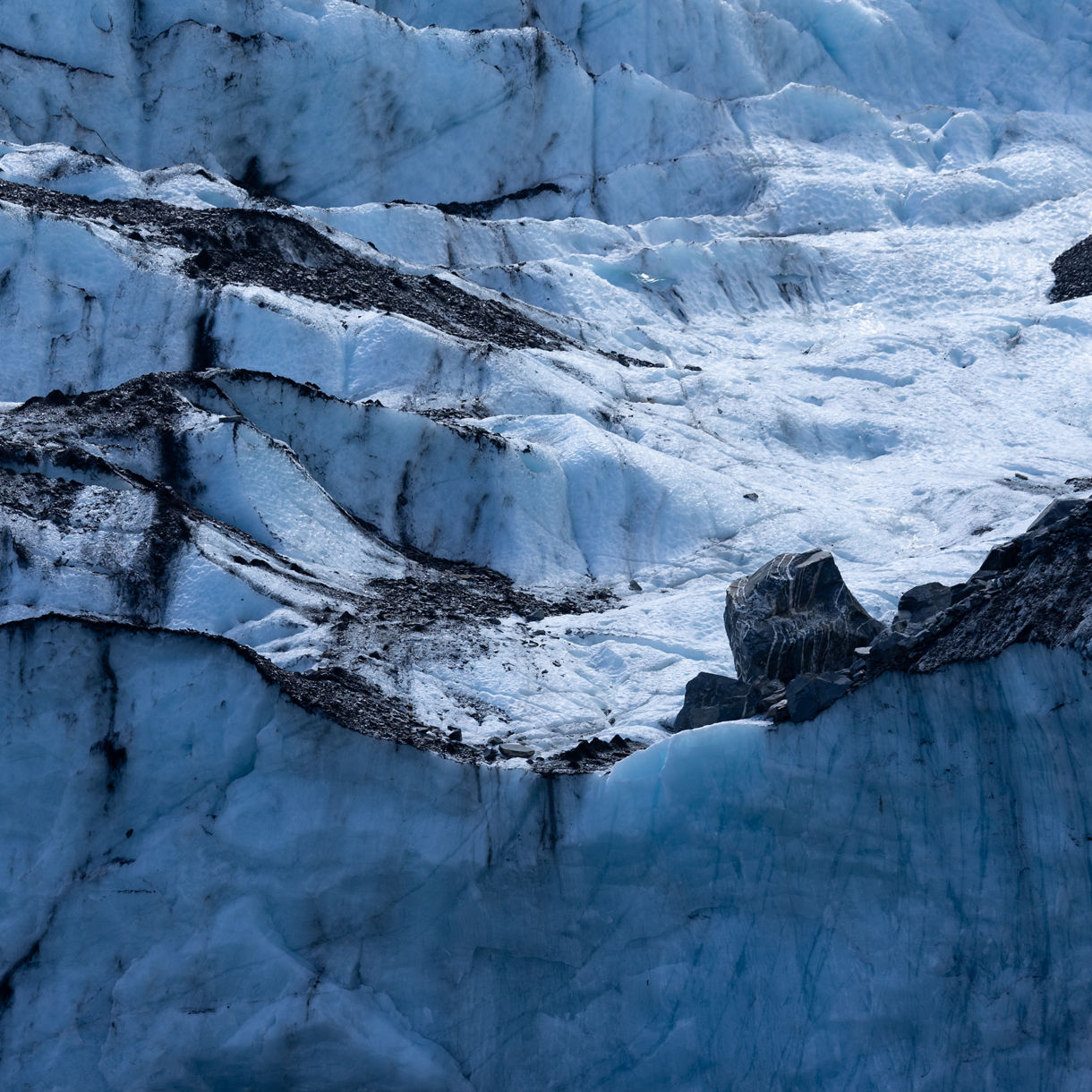 Close-up view of Portage Glacier's icy wall with deep blue crevasses, dark sediment layers, exposed rock, and floating ice chunks on the water's surface.