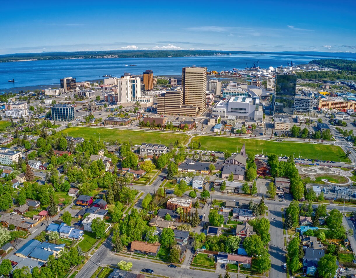 Aerial view of downtown Anchorage, Alaska on a clear summer day, showing modern buildings, green residential neighborhoods, and the blue waters of Cook Inlet in the background.