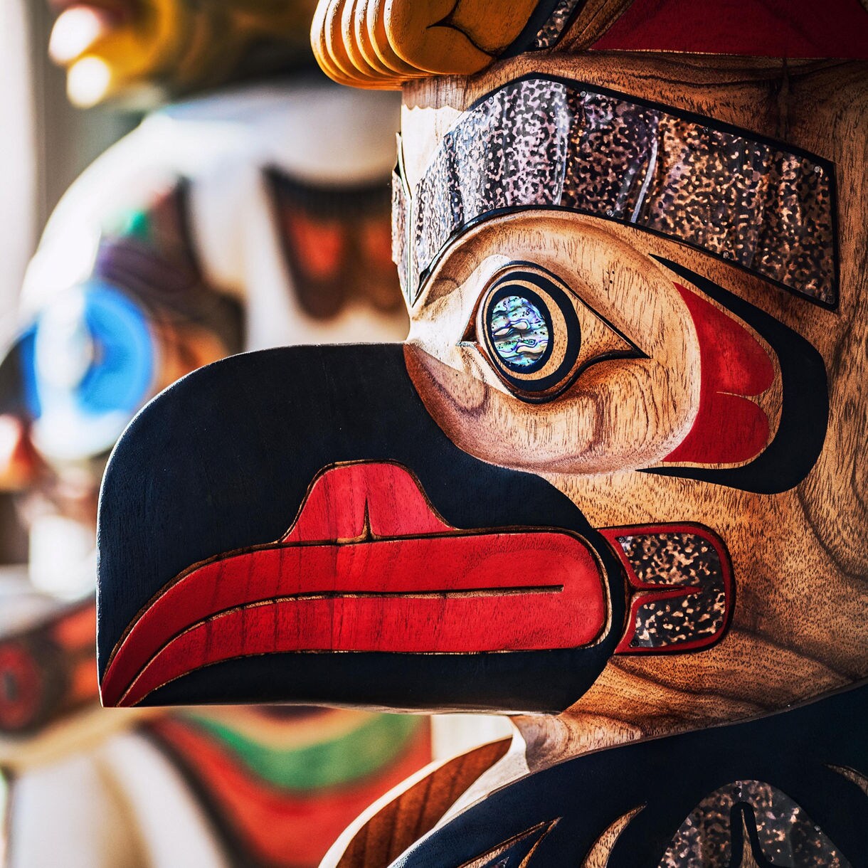 Close-up of a colorful Native Alaskan totem pole featuring a bird design with abalone shell inlays, surrounded by blurred totems in the background.