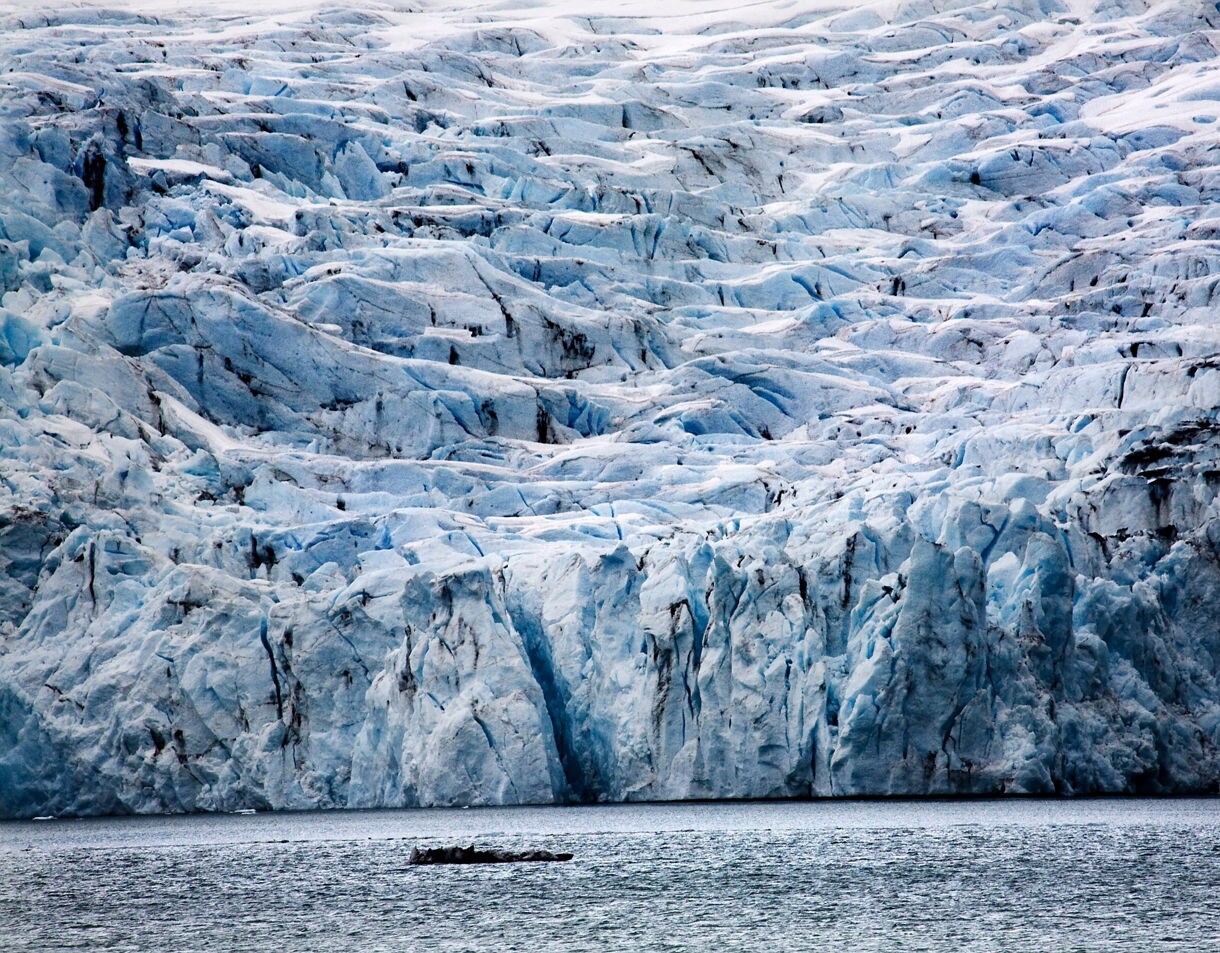 Massive blue and white ice formations of Portage Glacier rising above the water, with deep crevasses and dark striations visible in the frozen surface.