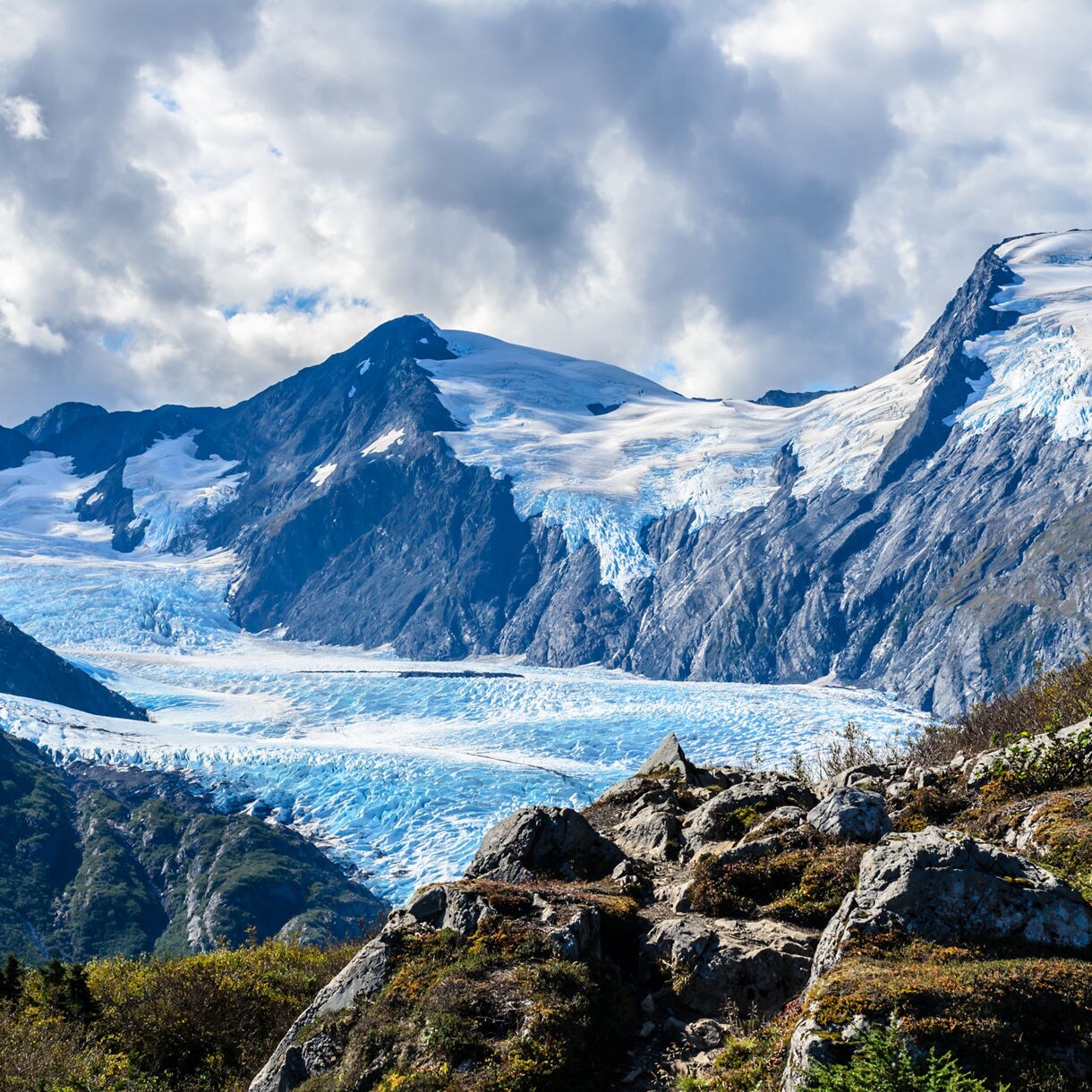 Expansive view of Portage Glacier in Alaska, with bright blue ice flowing through a mountain valley surrounded by rocky slopes and patches of greenery under a cloudy sky.