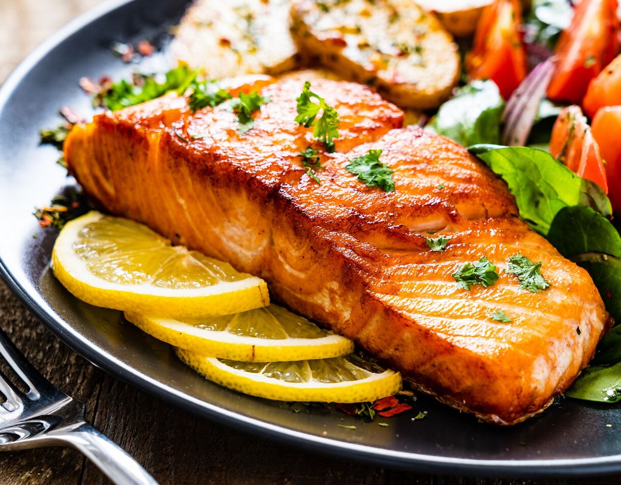 Close-up of a grilled salmon fillet garnished with parsley, plated with lemon slices, leafy greens, roasted potatoes, and fresh tomato salad.