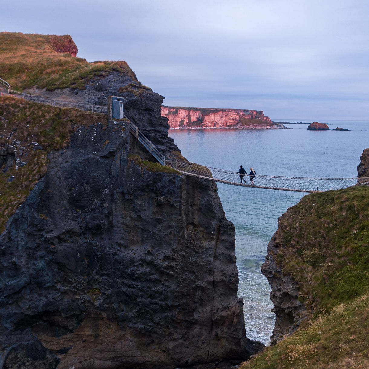Two people walk across a narrow rope bridge suspended between steep rocky cliffs above the ocean. The scene is set along a rugged coastline with grassy paths winding over the cliff tops. Soft, overcast light and muted colors create a calm, slightly dramatic atmosphere. Distant cliffs and waves below emphasize the height and adventurous nature of the crossing.