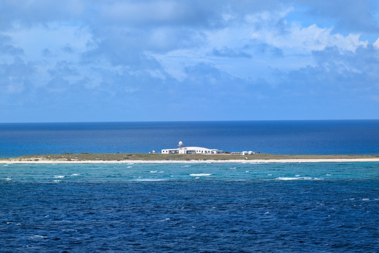 Small white research buildings on a flat, narrow coral island surrounded by turquoise water and deep ocean.
