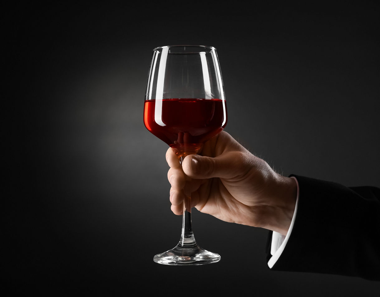 Hand in a black suit holding a glass of red wine against a dark background.