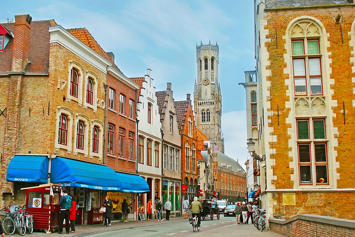 Street view of Wollestraat in Bruges, Belgium, with brick buildings, shops and the medieval Belfry of Bruges rising in the background.