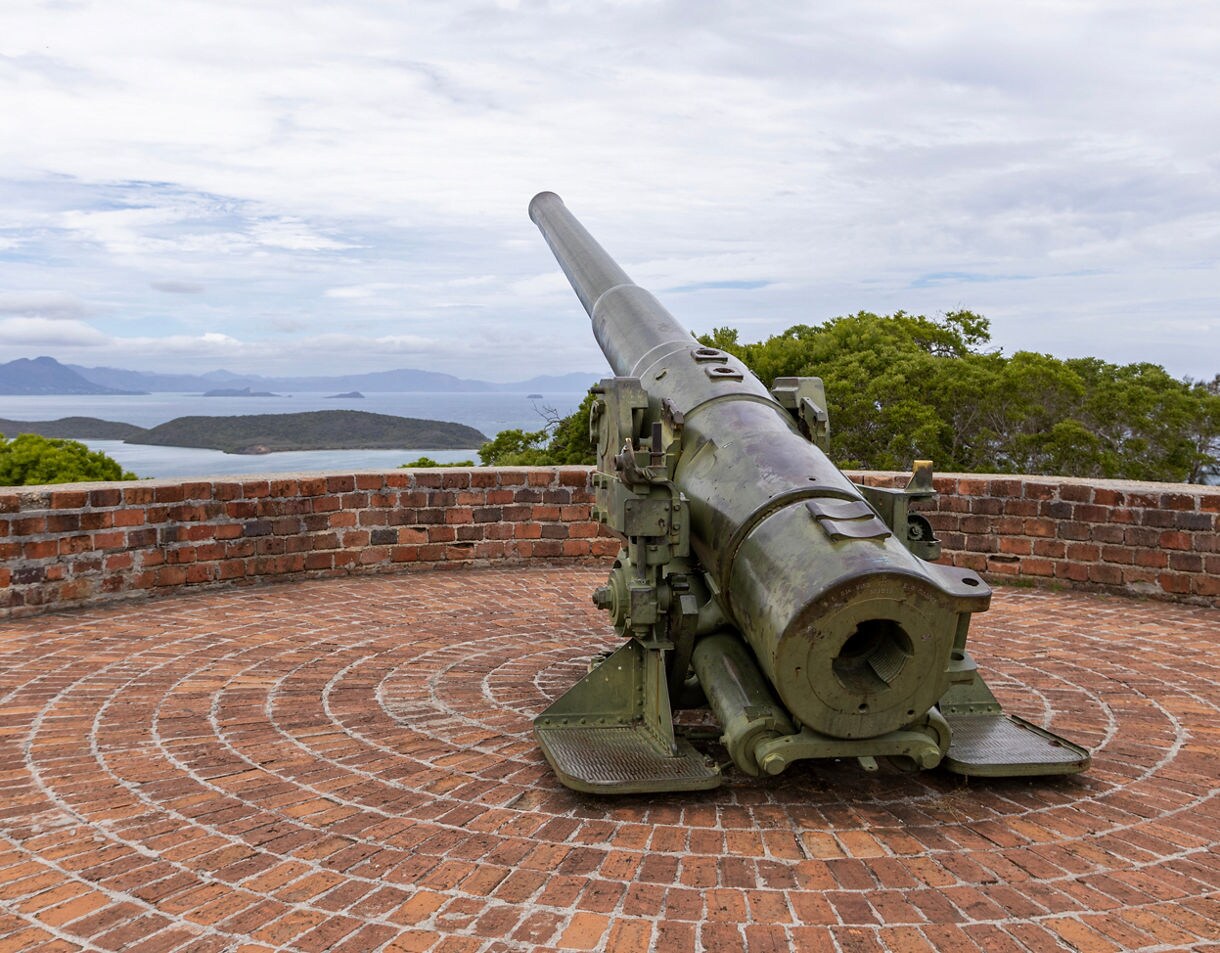 Old World War II coastal artillery gun mounted on a circular brick platform overlooking blue ocean waters and small green islands under a cloudy sky.