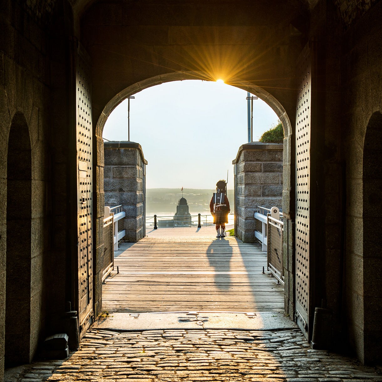 A person walks through a historic stone archway illuminated by warm sunlight. The setting appears to be a coastal fort or castle, with cobblestone flooring and wooden gates. The silhouette of the individual and the golden light create a serene and timeless atmosphere.