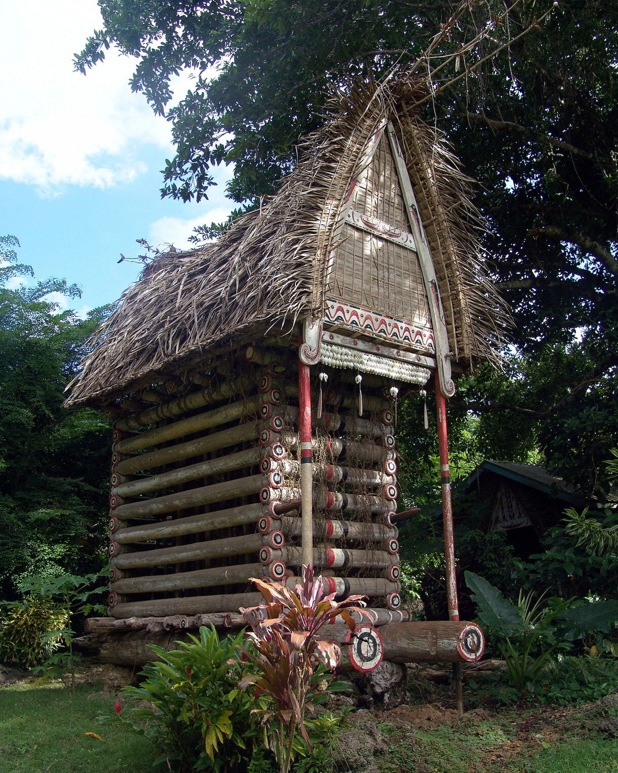 Elevated wooden yam house built from stacked logs with intricate red, white and black painted patterns, topped by a steep thatched roof and surrounded by dense tropical greenery.