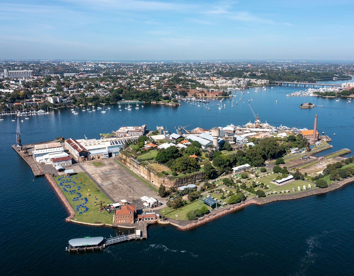 Aerial view of Cockatoo Island featuring industrial buildings, historic sandstone structures, open lawns, and docks surrounded by the blue waters of Sydney Harbour with the cityscape in the background.