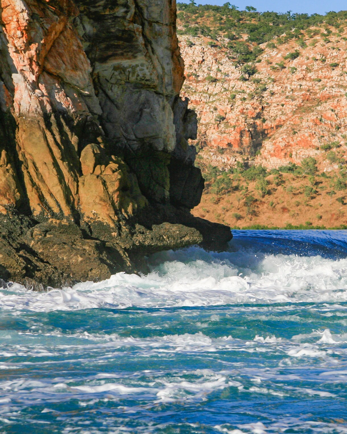 Close-up view of strong tidal currents pushing through a narrow gap between tall orange sandstone cliffs, with whitewater swirling in the foreground and rocky hills in the background.