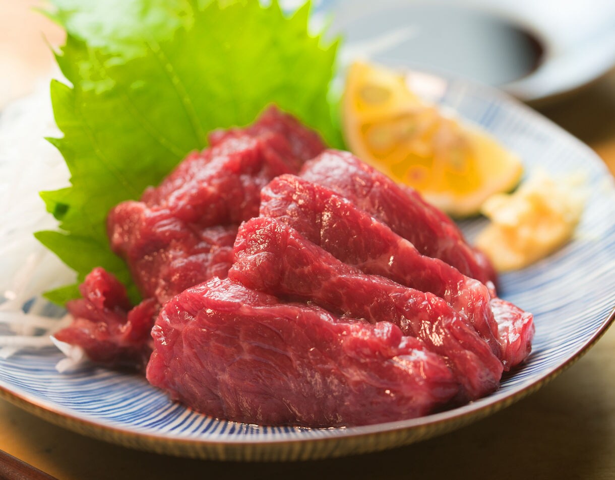 Close-up of fresh red basashi sashimi arranged on a blue plate with shredded daikon, shiso leaves, lemon, and a small portion of grated ginger.
