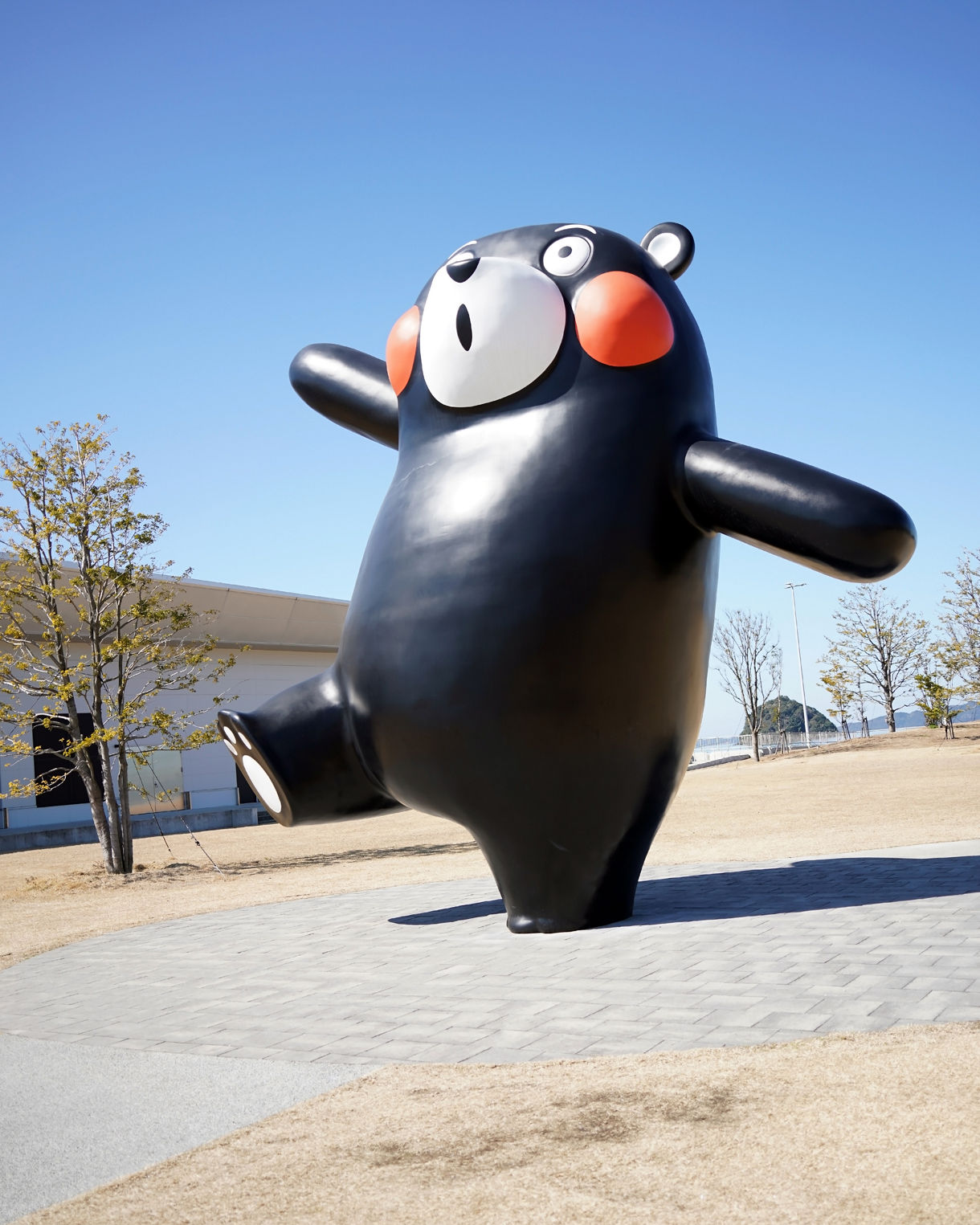 A large outdoor statue of Kumamon, the black bear mascot with red cheeks, stands playfully on one leg in an open plaza with trees and clear skies in the background.