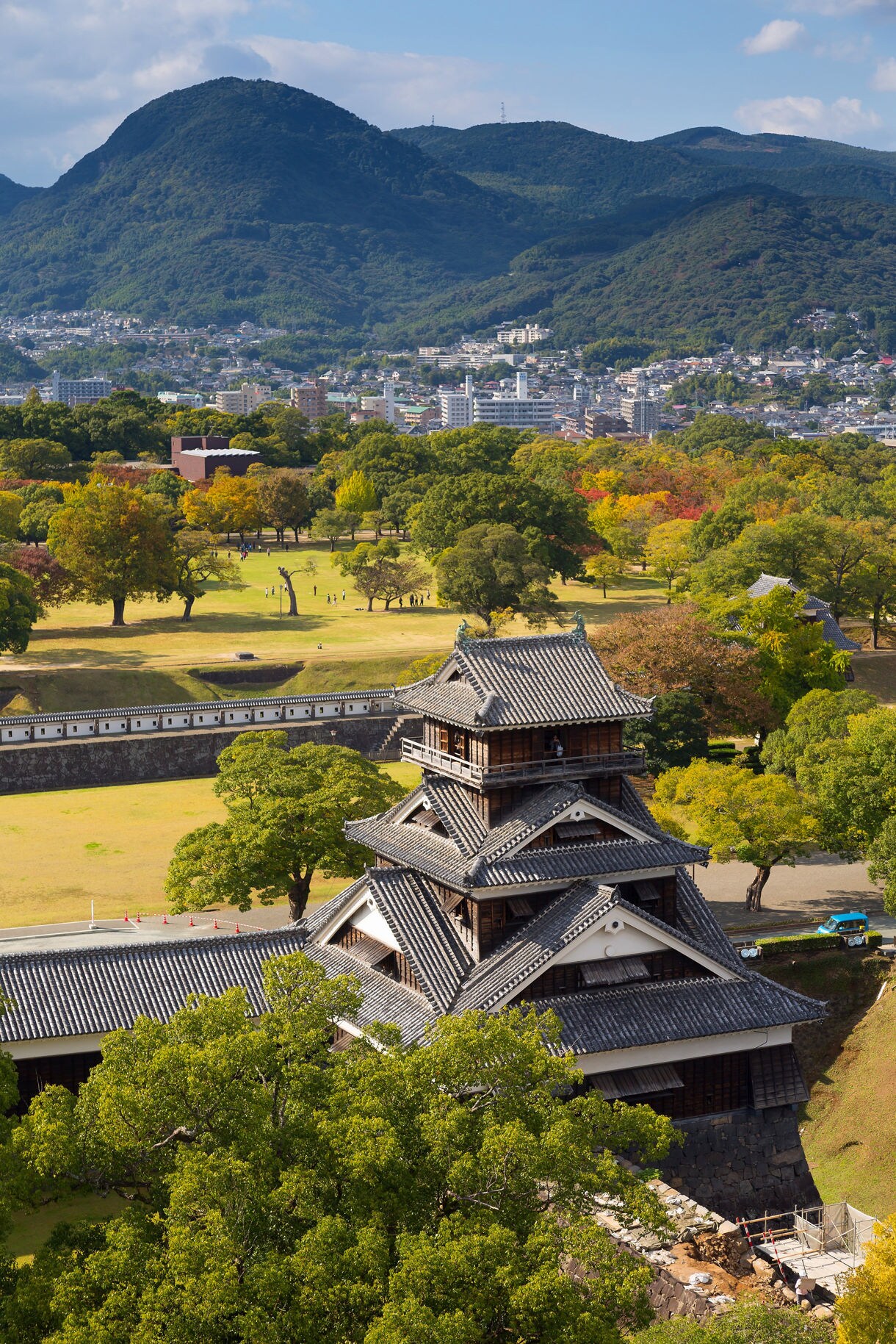 Elevated view of Kumamoto Castle’s wooden and tile-roofed tower surrounded by lush trees, with a distant city and forested mountains under a clear sky