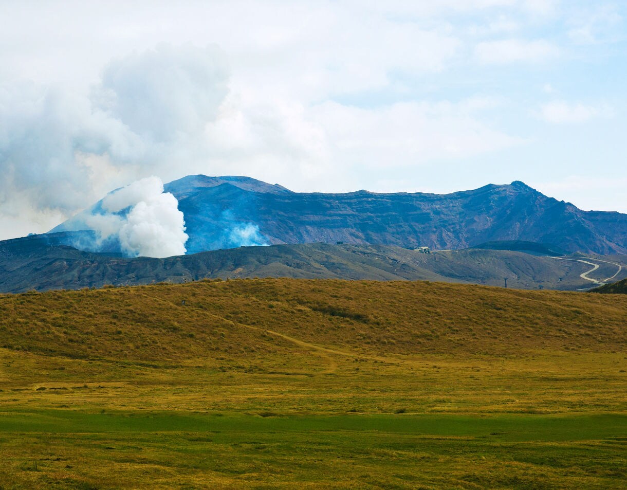 A smoking volcanic crater on Mount Aso sends white plumes into the sky above dark rocky slopes, with golden grasslands stretching across the foreground.