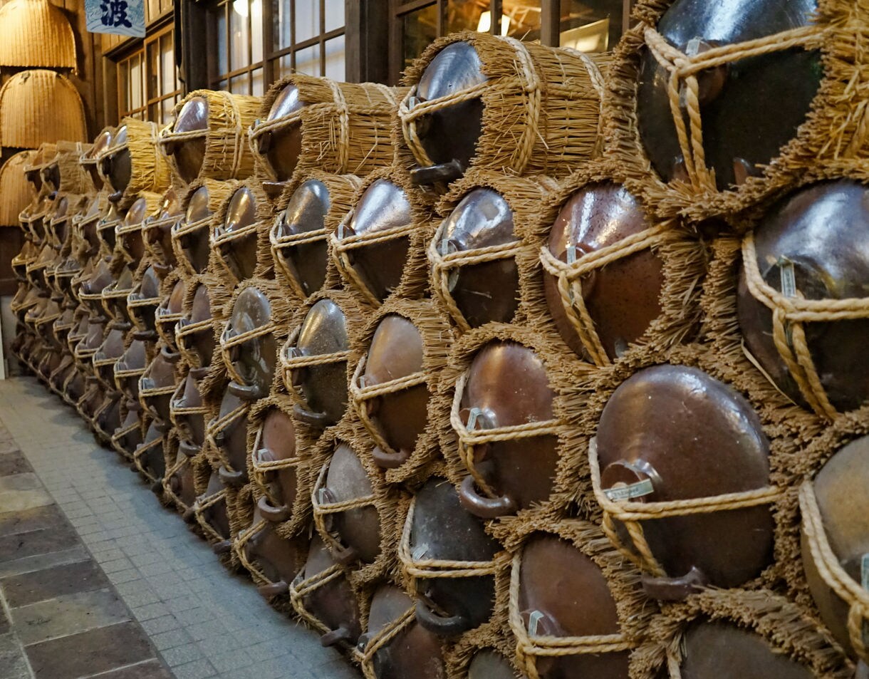 Dozens of large clay fermentation jars wrapped in thick straw mats are stacked neatly along a hallway inside a shochu brewery.