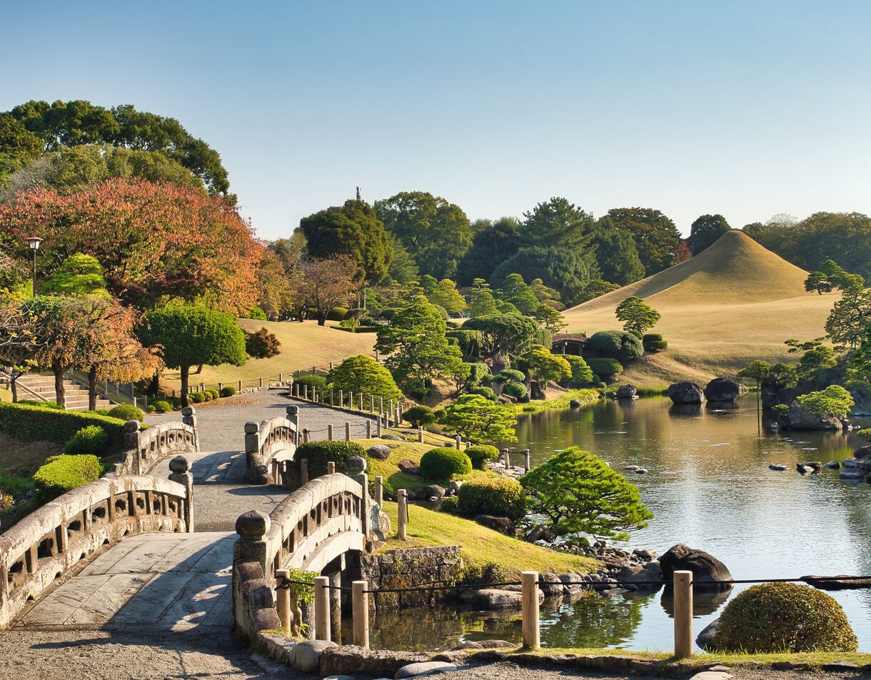 A traditional Japanese garden featuring arched stone bridges, manicured trees, a reflective pond, and a smooth conical hill under clear blue skies.
