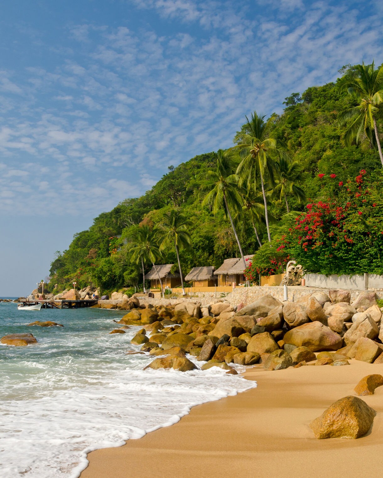 Palm trees, stone seawall and rustic beach huts line the sandy shore of Yelapa near Puerto Vallarta with waves rolling in.
