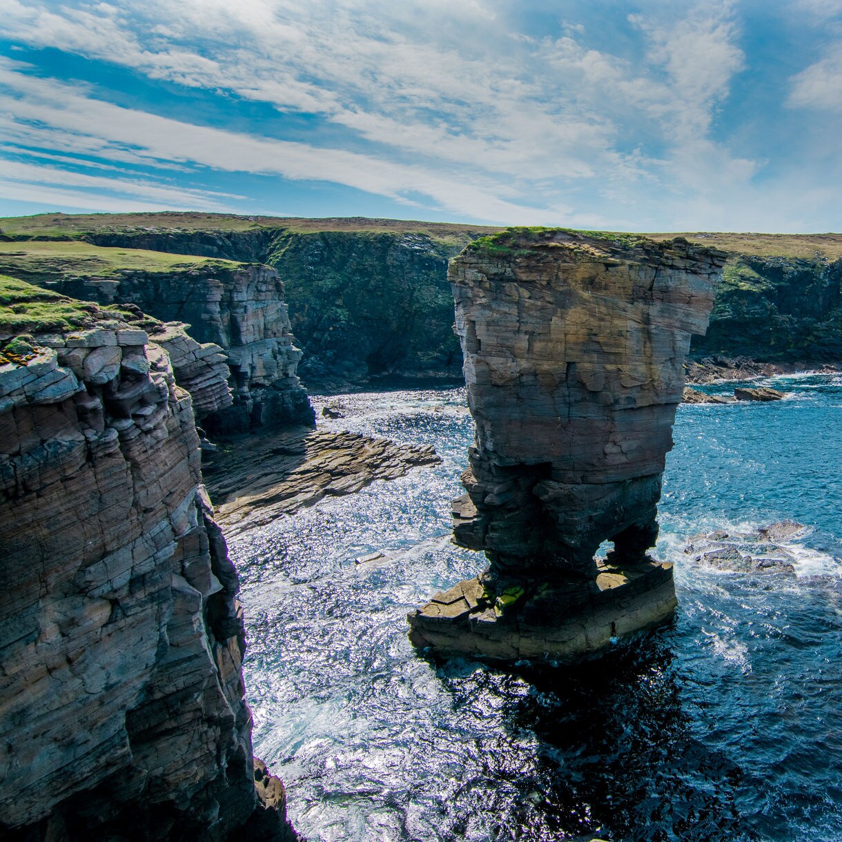 Towering sea stack surrounded by blue waters at Yesnaby Cliffs, with rocky shoreline and grassy cliffs in the distance under a partly cloudy sky.