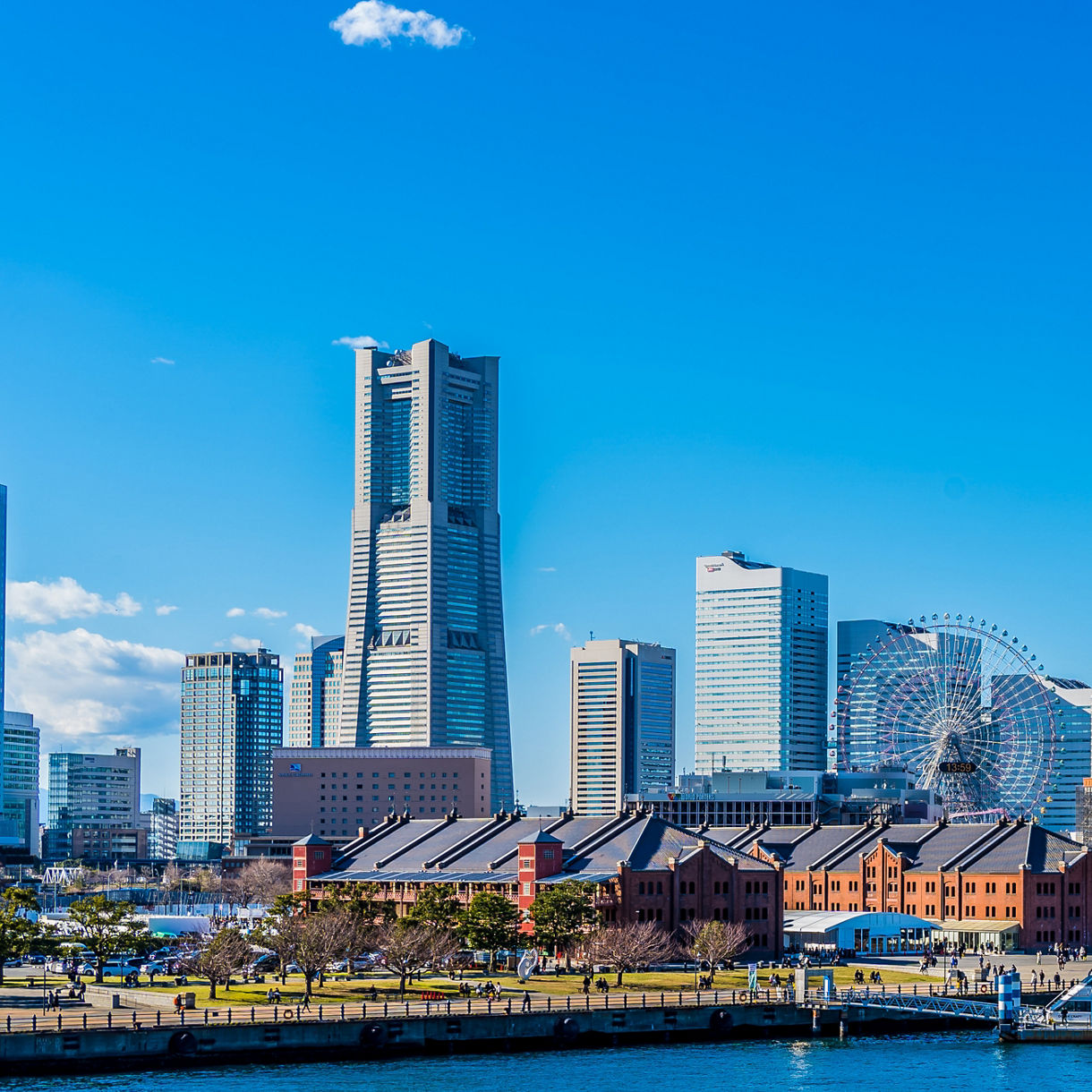 The Yokohama waterfront skyline featuring the Landmark Tower, a Ferris wheel, historic red brick warehouses and a harbor with docked boats under a clear blue sky.