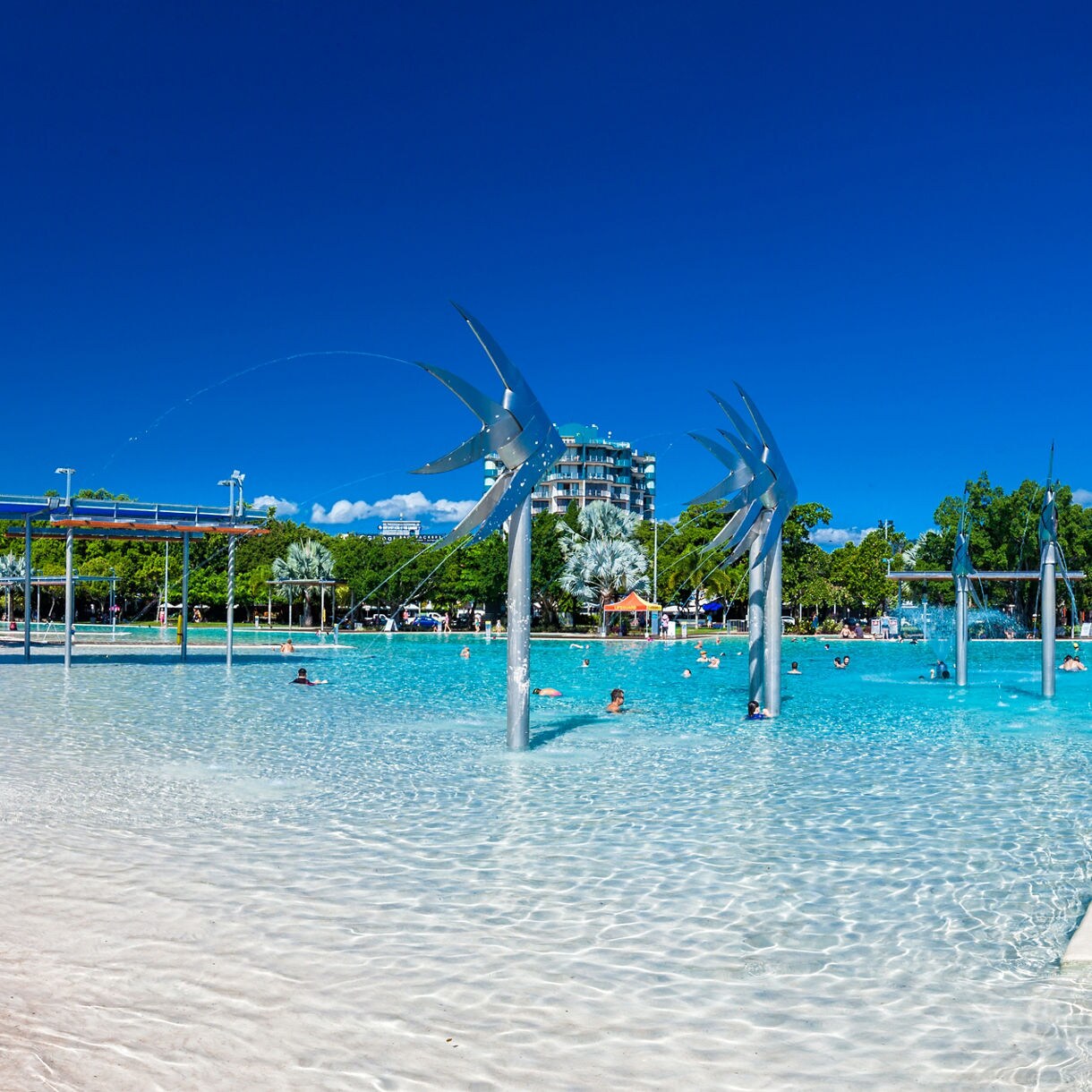 A shallow swimming lagoon beside a sandy beach with silver palm trees, metal fish sculptures and people relaxing and swimming, set along the Cairns waterfront under a clear blue sky.
