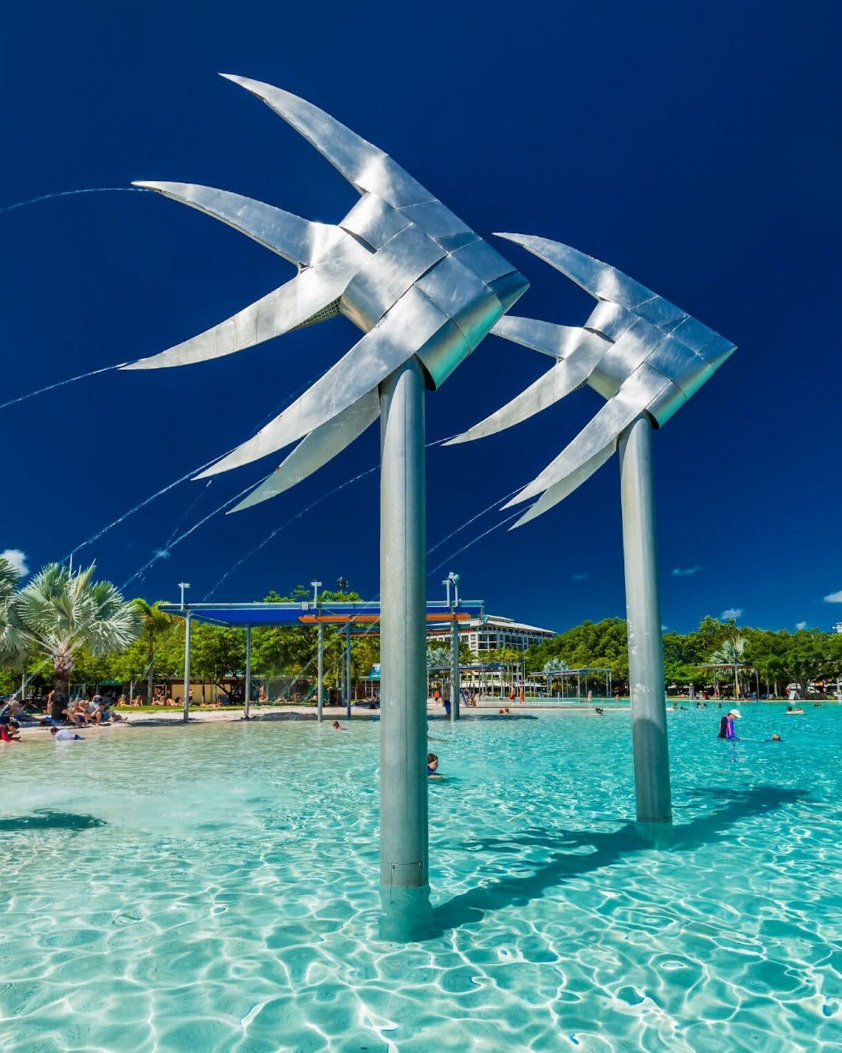 Large metal fish sculptures spouting water over a shallow turquoise swimming lagoon, with people relaxing in the clear water and trees lining the background under a deep blue sky.