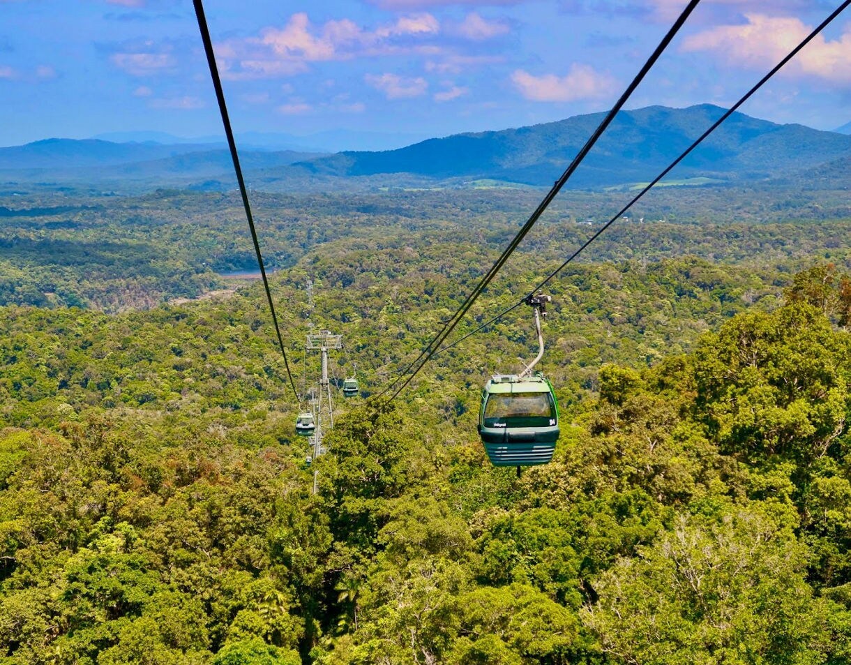 Green cable cars suspended above a vast rainforest landscape with rolling hills and blue mountains in the distance under a bright sky.