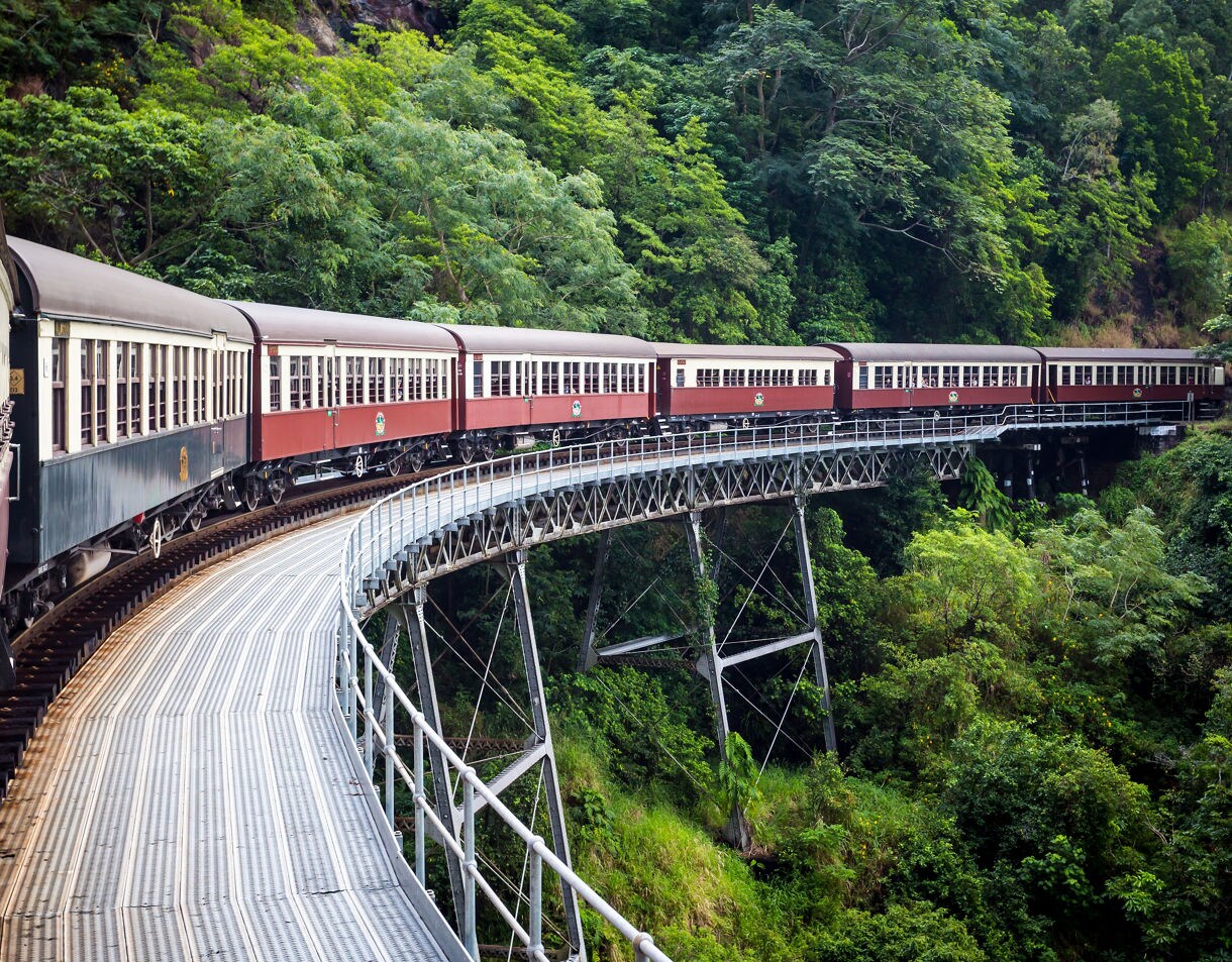 A vintage red-and-cream train traveling along a curved steel bridge through dense green rainforest, seen from one of the train cars.