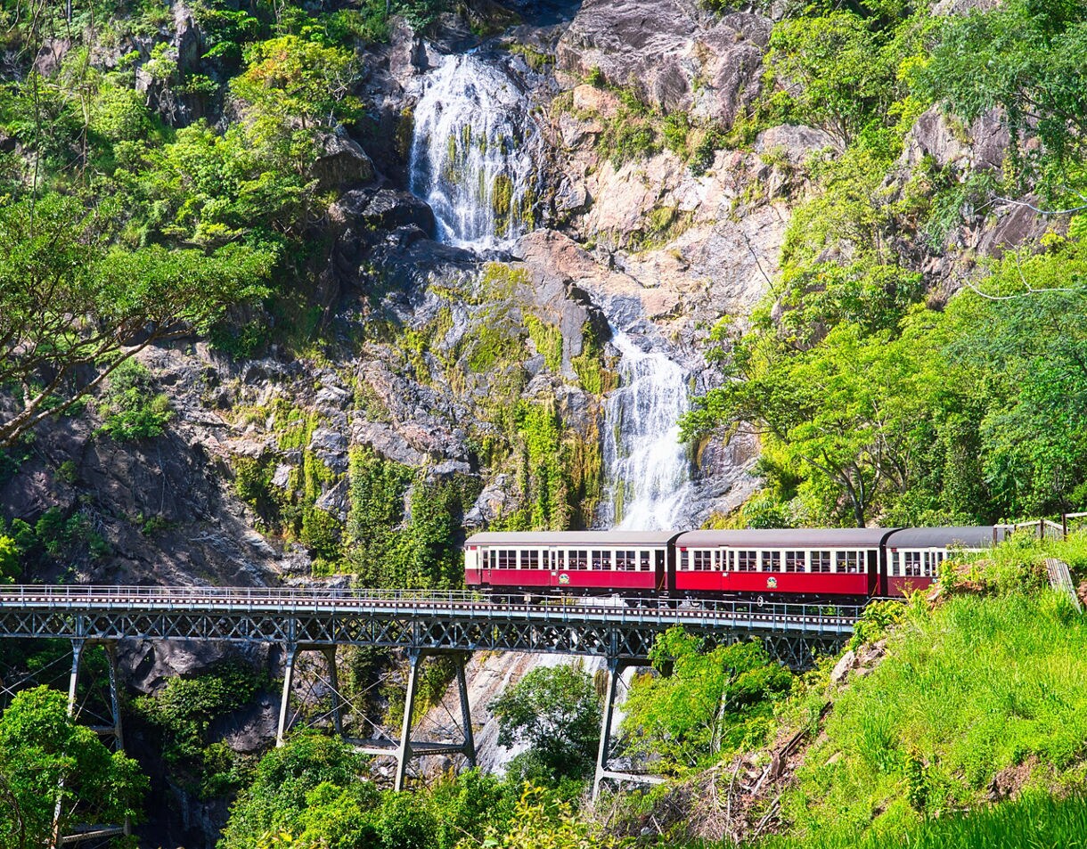 A vintage red-and-cream train crossing an elevated railway bridge in a tropical mountain landscape, with a cascading waterfall flowing down rocky cliffs behind it.
