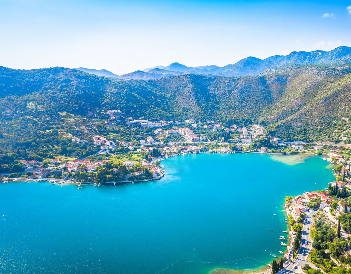 Aerial view of Zaton Bay in Croatia, featuring calm turquoise water surrounded by green hills and small coastal villages with red-roofed houses.