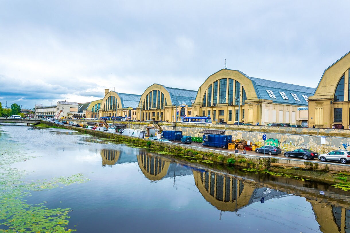 Exterior view of Riga Central Market in Latvia, housed in massive former zeppelin hangars with arched windows, reflected in the canal below on a cloudy day.