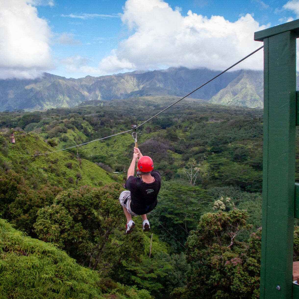 Behind-the-back view of a person wearing a red helmet riding a zipline over a deep green valley filled with thick trees and tropical foliage, with dramatic mountain peaks under white clouds in the background.