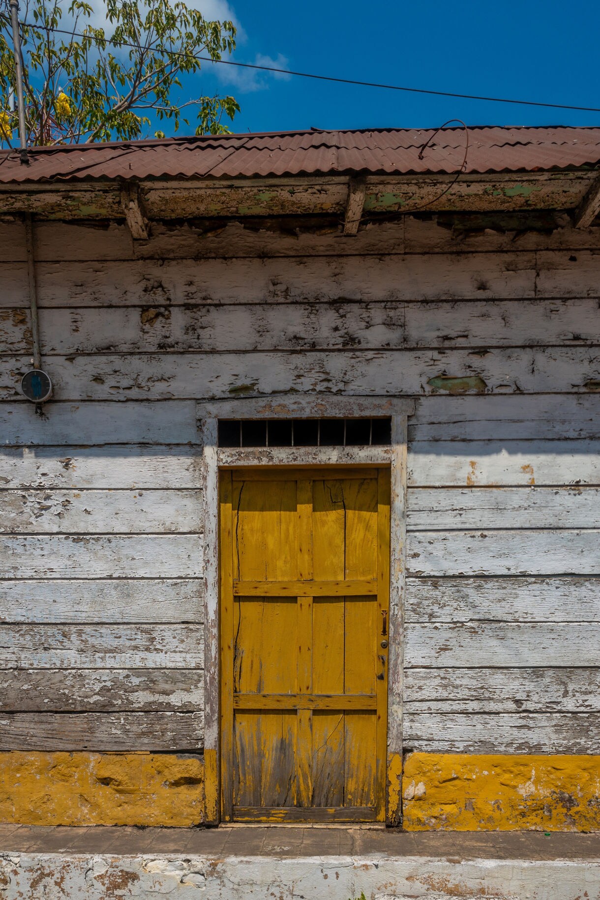Aged wooden building with peeling white paint and a bright yellow door, set beneath a corrugated metal roof under a clear blue sky.