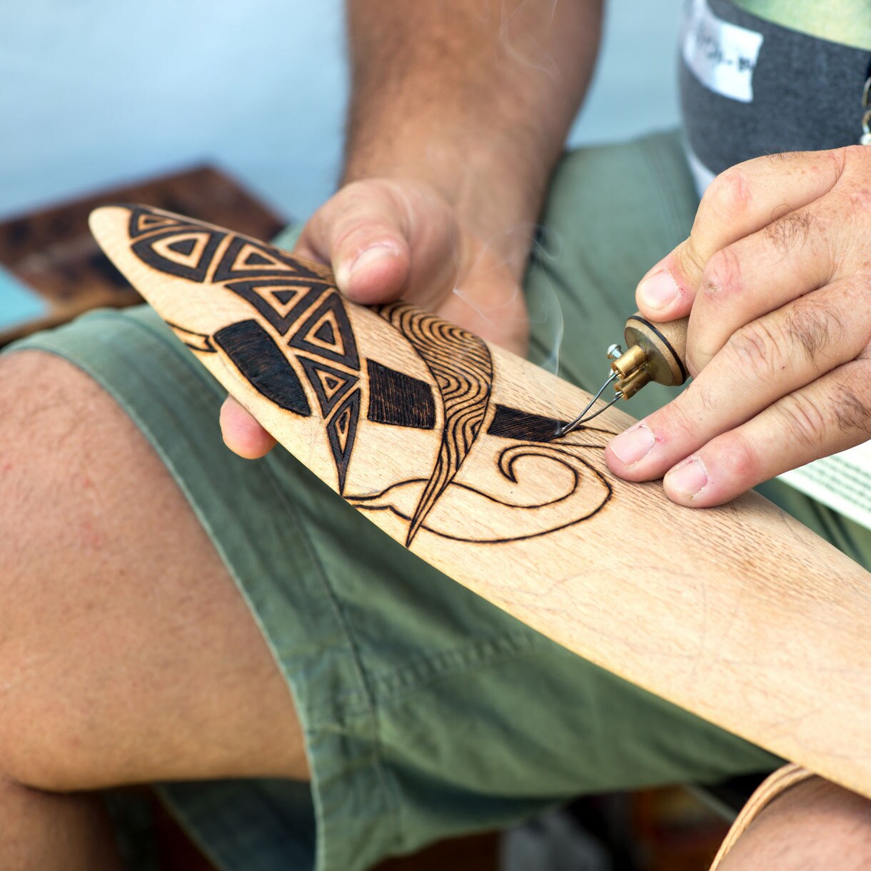 Close-up of hands using a woodburning tool to create Aboriginal-style designs on a wooden boomerang.