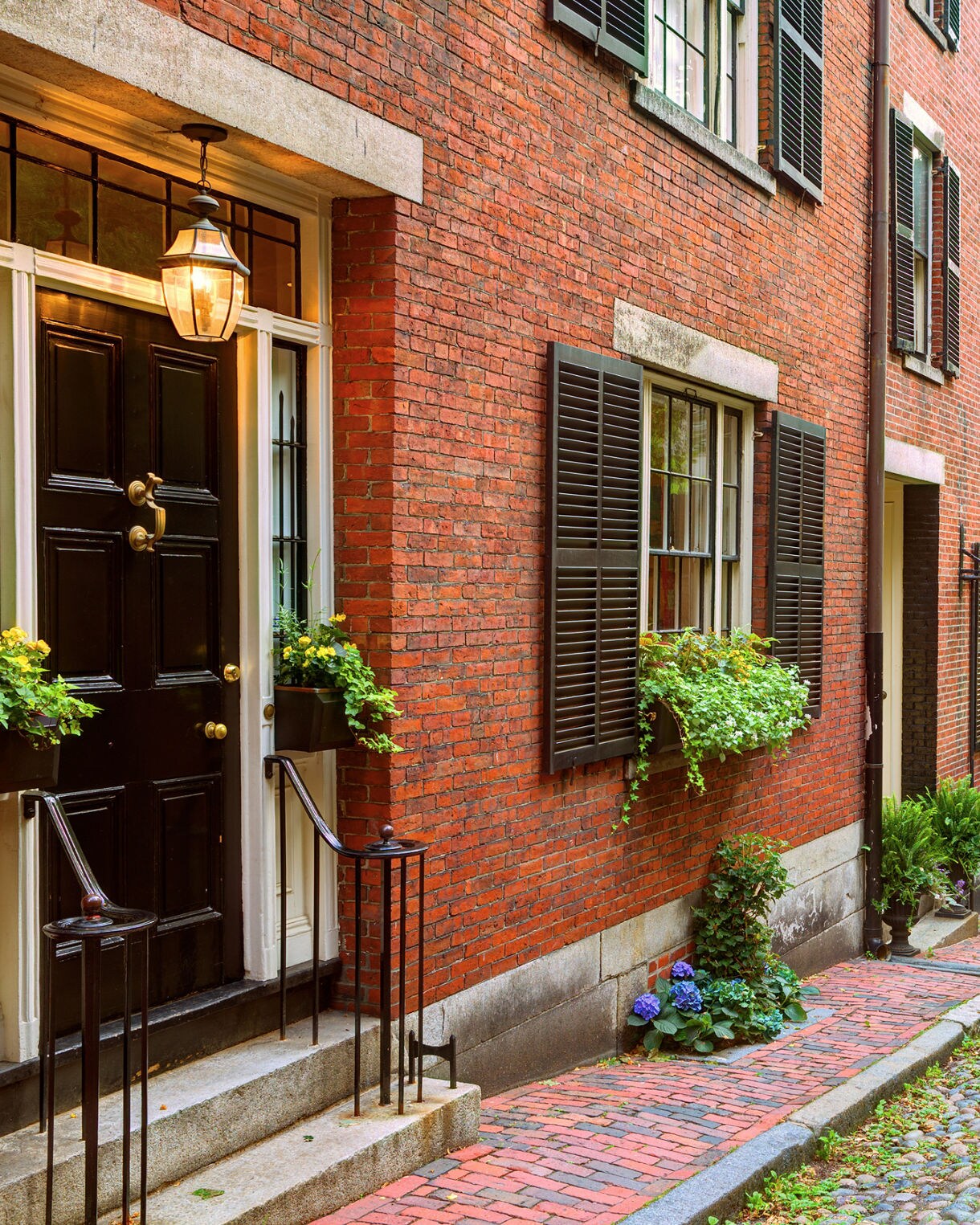 Narrow cobblestone street lined with historic red-brick row houses, black shutters, hanging plants and an American flag in Boston’s Beacon Hill neighborhood.