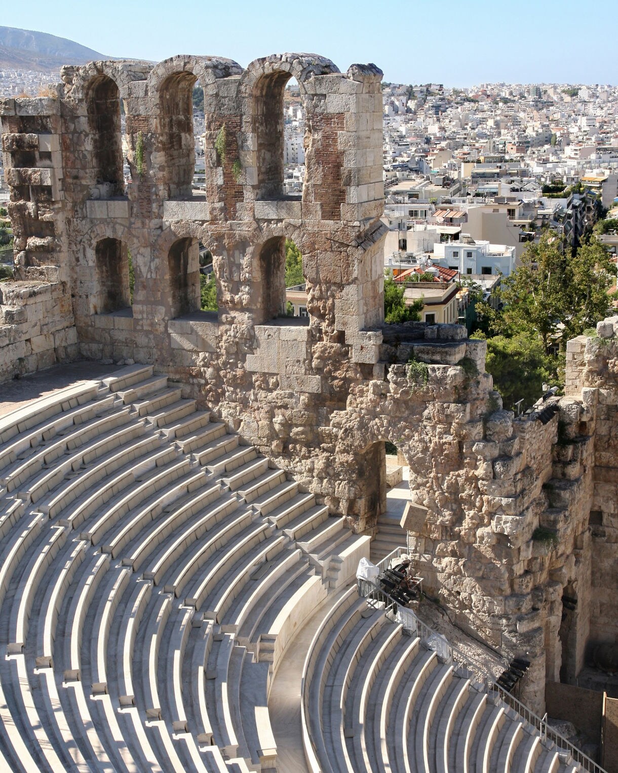 View of a semicircular stone amphitheater with tiered marble seating and a partially preserved stone wall overlooking the city of Athens.