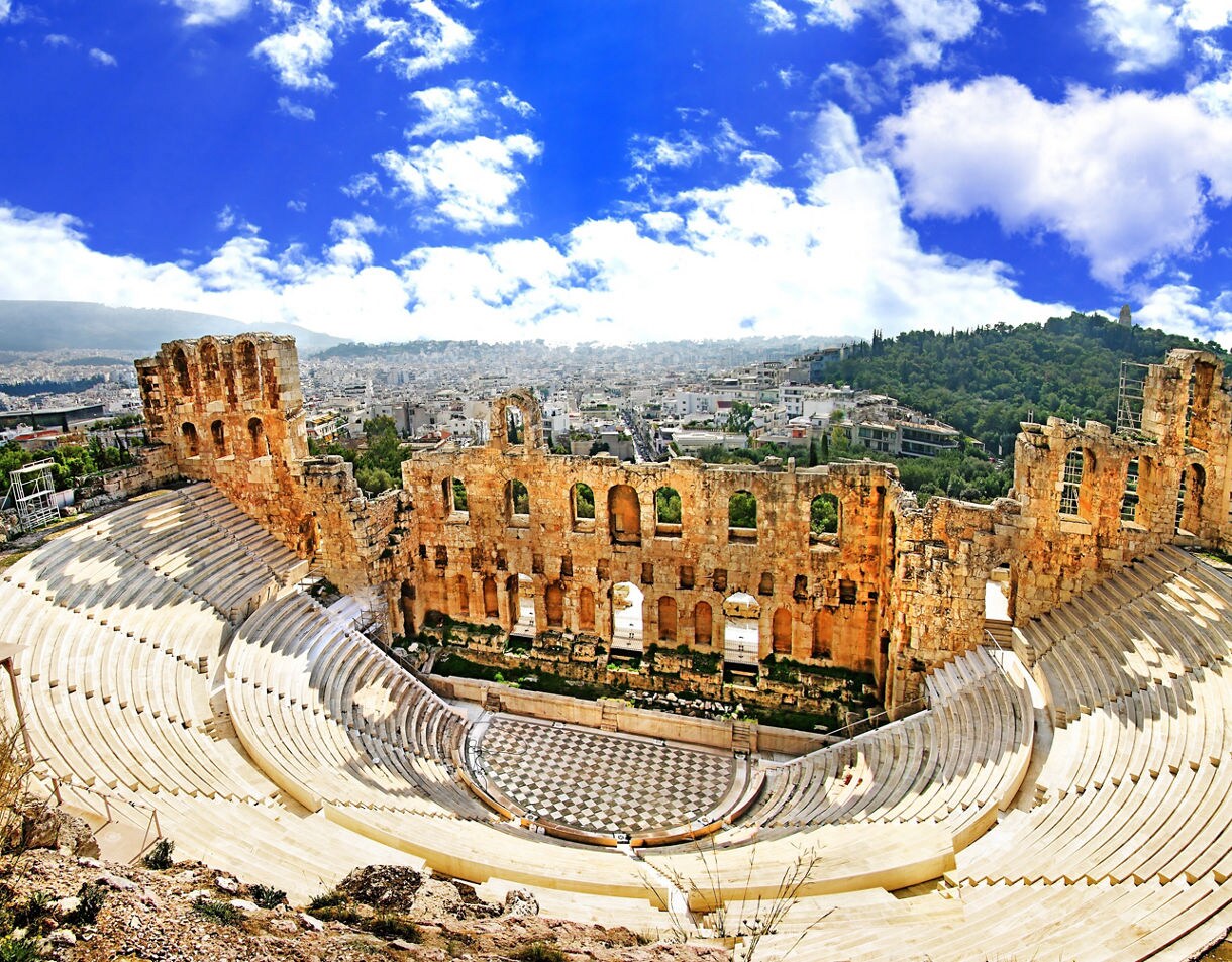 Panoramic view of the ancient Odeon of Herodes Atticus amphitheater in Athens, with stone seating curving around a central stage and the modern city spreading out behind it under a bright blue