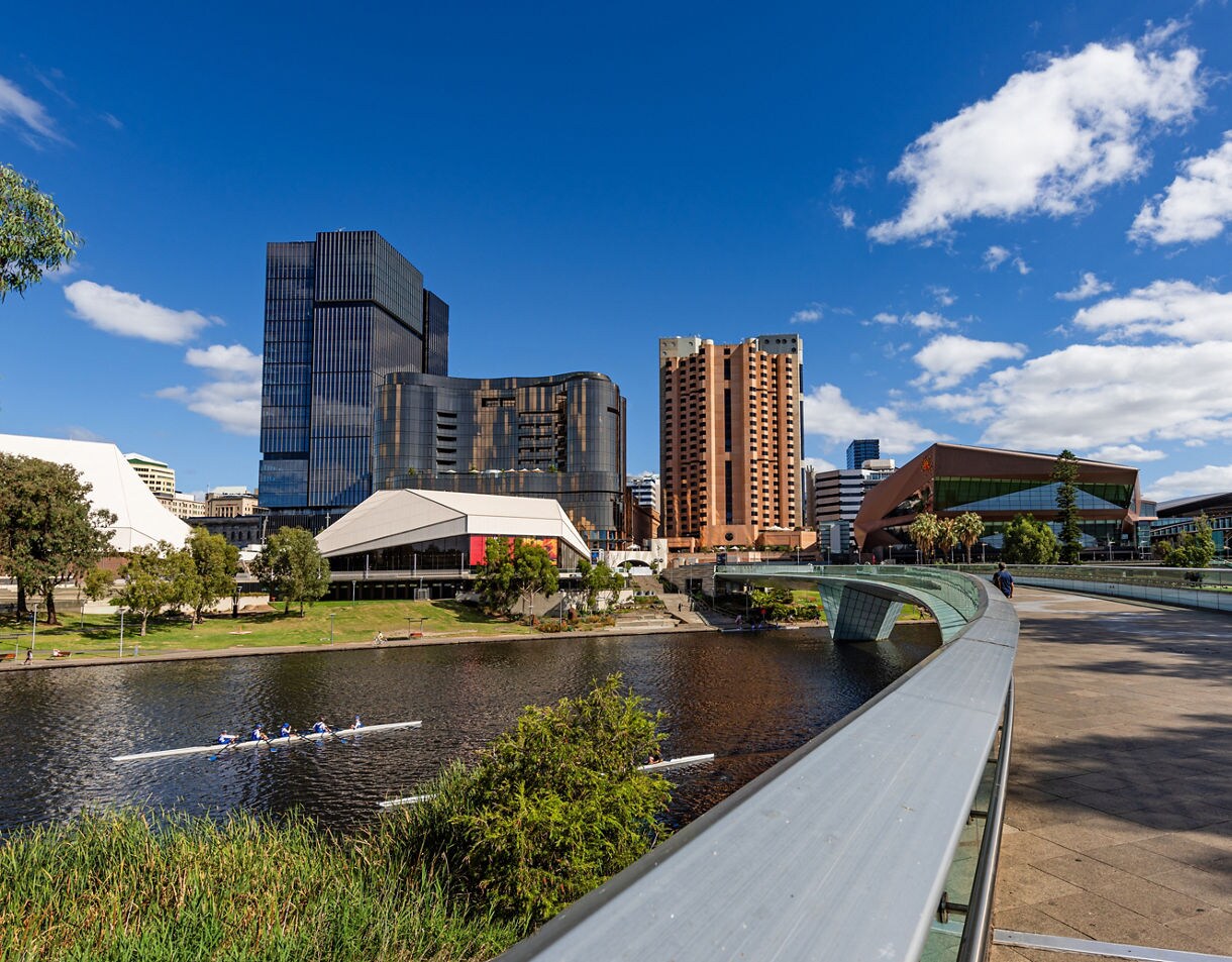 Modern buildings and a pedestrian bridge along the River Torrens with rowers on the water under a bright blue sky.
