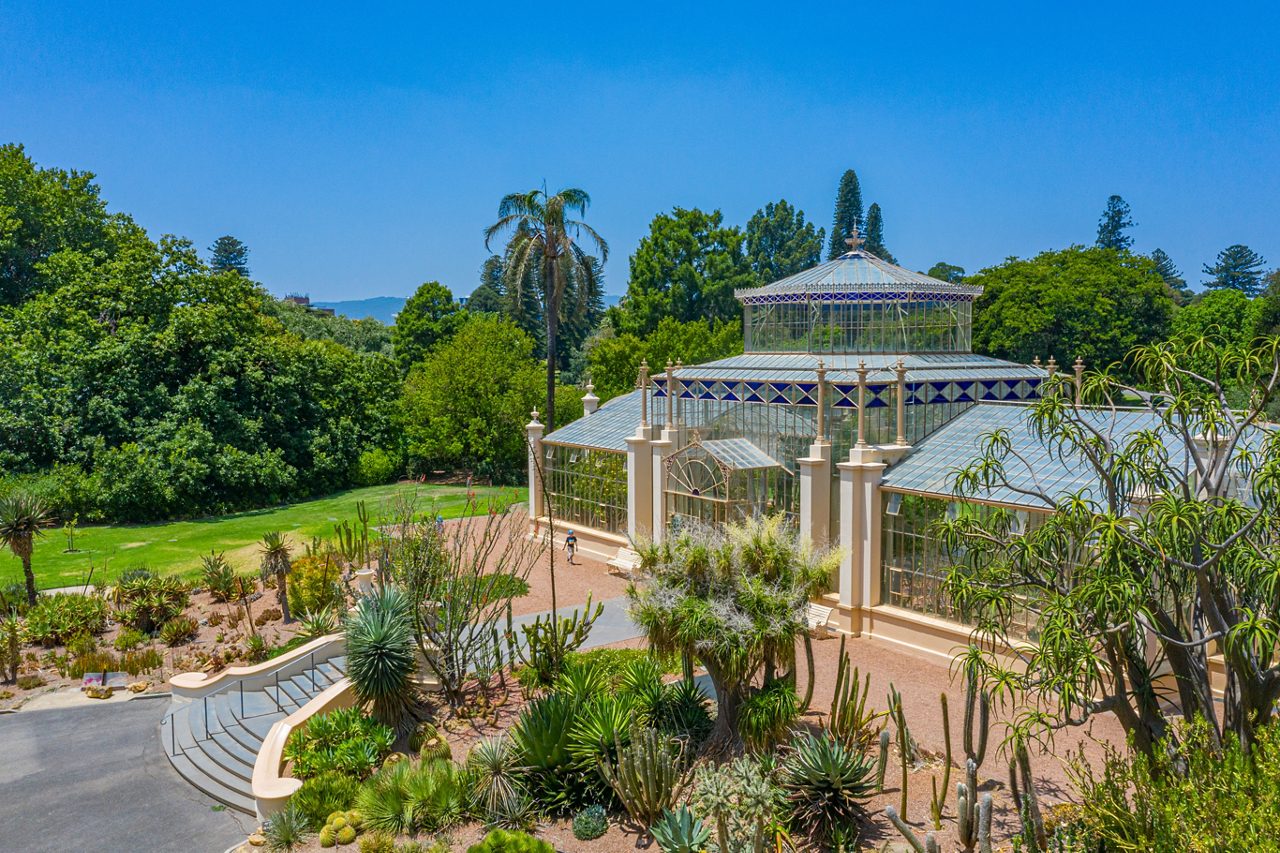 A large ornate glasshouse set among lush trees and desert plants in a botanical garden under a bright blue sky.