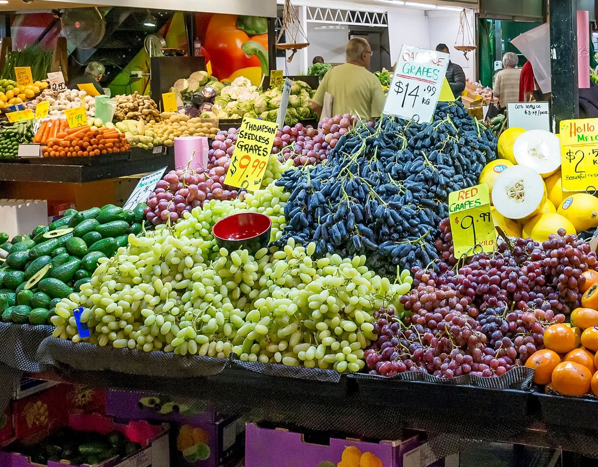 A colorful market stall displaying piles of grapes, avocados, persimmons and other fresh fruits and vegetables with handwritten price signs.