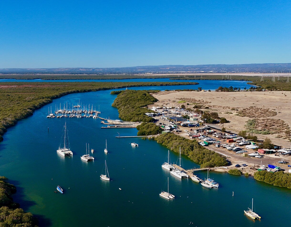 Aerial view of a narrow blue inlet lined with mangroves, sailboats anchored in the water and a small marina on the right, with flat countryside in the distance.