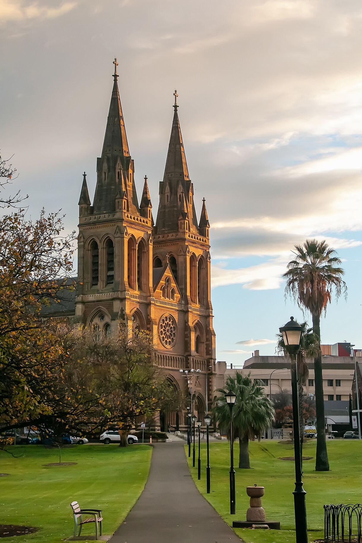 A tall sandstone cathedral with twin spires glowing in warm evening light, seen from a garden pathway lined with lampposts and palm trees.