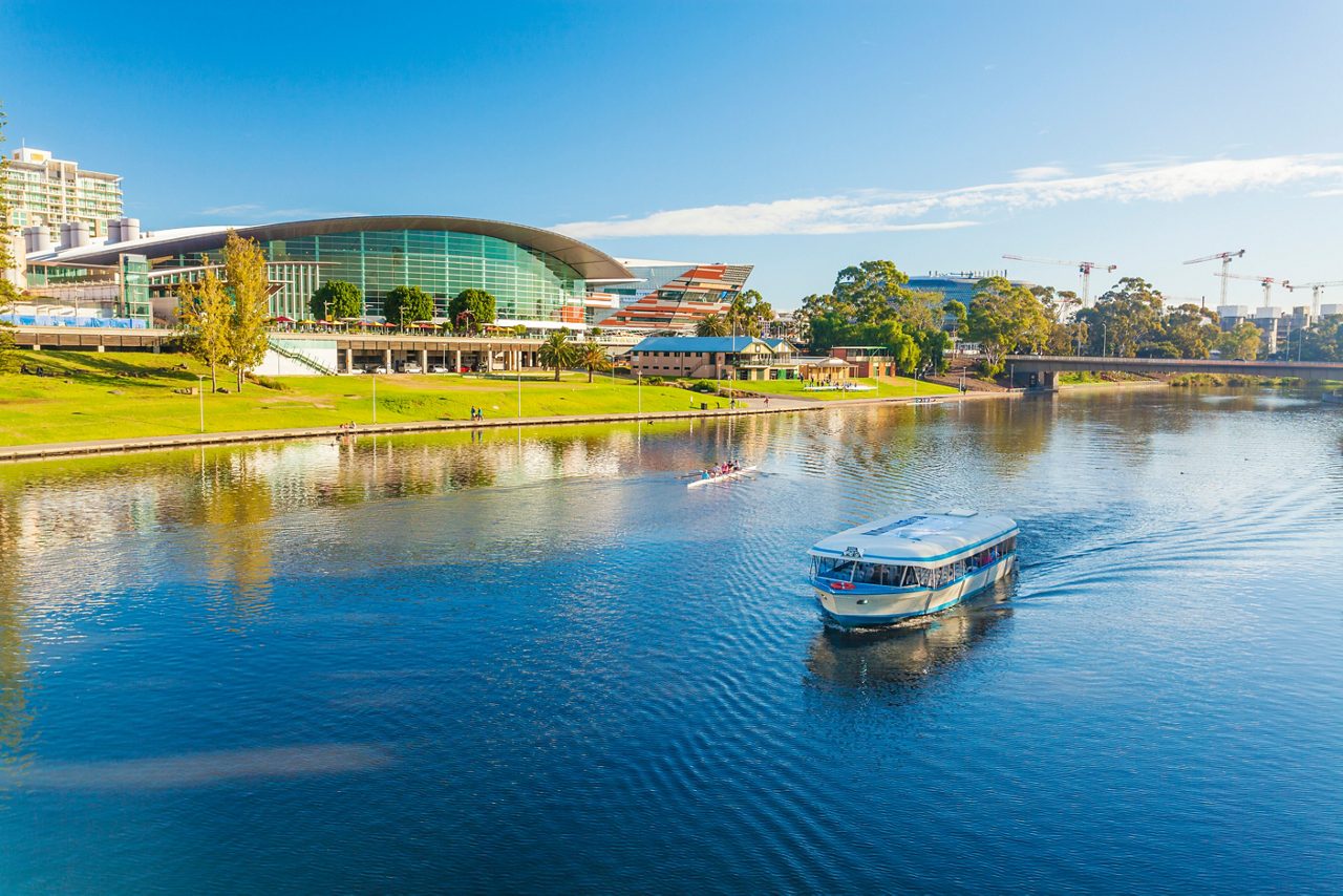 A sightseeing boat glides along a wide river with a modern glass building, green lawns and trees on the opposite bank under a bright blue sky.