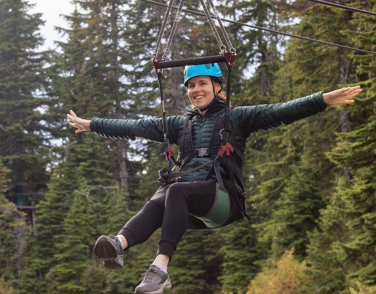 A smiling woman wearing a blue helmet and safety harness rides a zipline with evergreen trees in the background.
